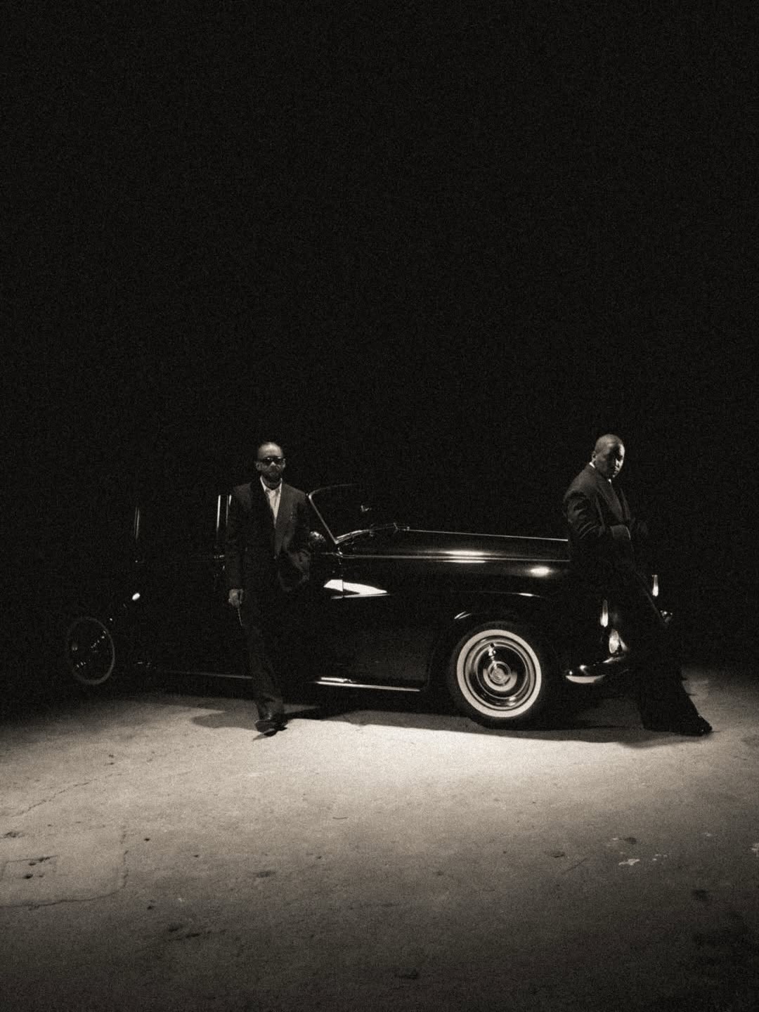 Two men standing beside a classic Rolls-Royce on a monochrome-lit set.