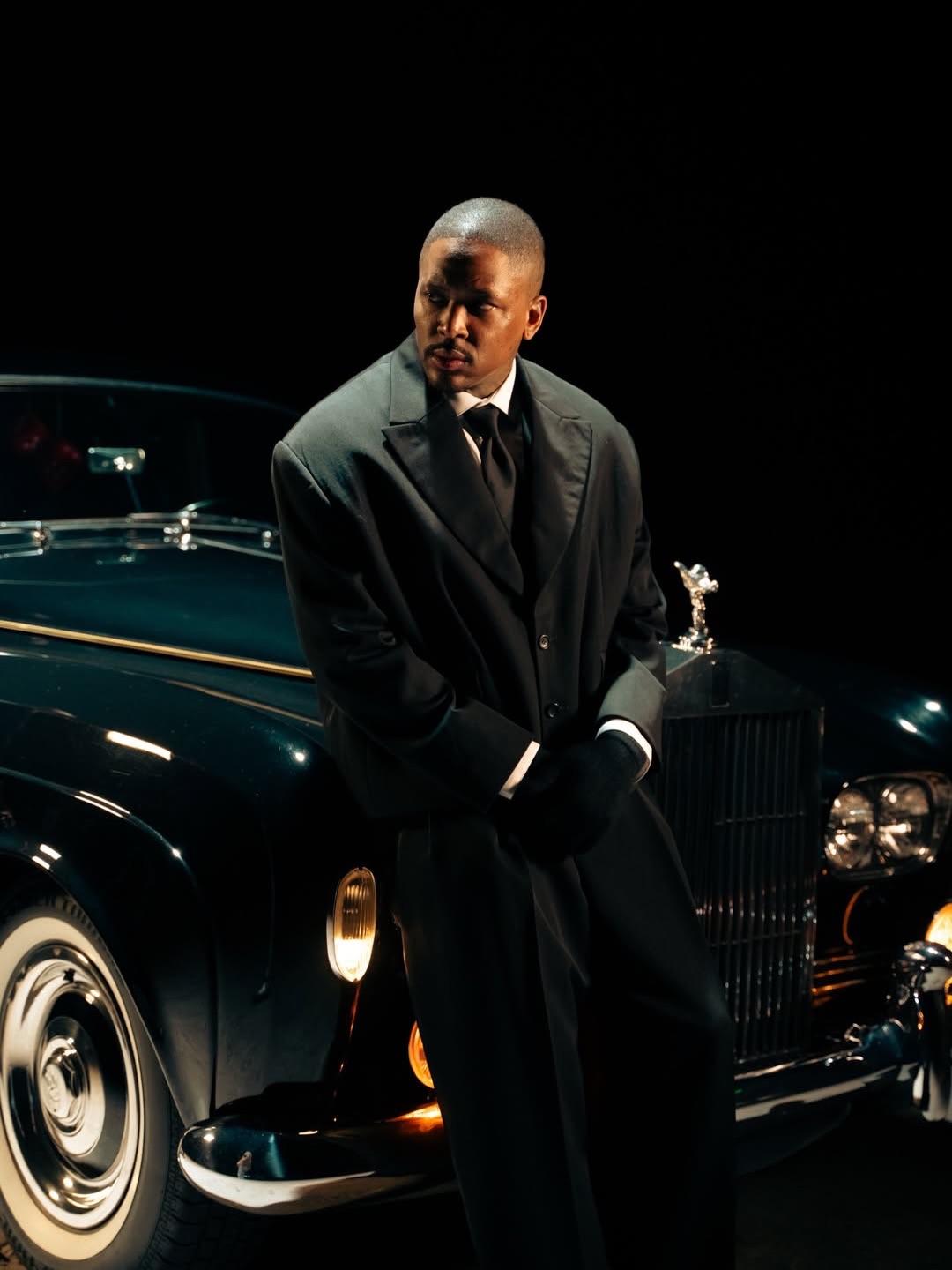 A man in a suit posing beside a classic Rolls-Royce under studio lighting.